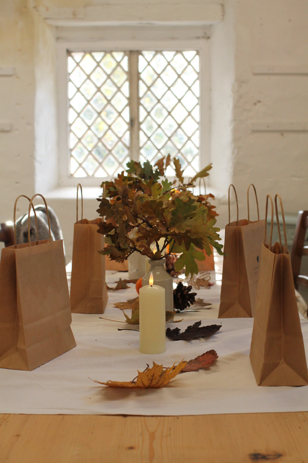 Brown paper gift bags with wire handles arranged on a white table runner in a bright room with a window and autumn leaf decorations.